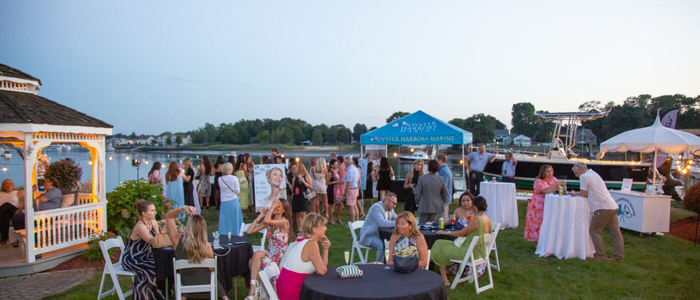 Guests at cocktail tables on the waterfront lawn at Danversport at dusk, with a gazebo, string lights, vendor tents, and boats in the marina behind them.