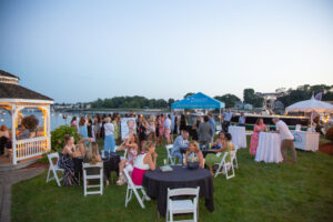 Guests at cocktail tables on the waterfront lawn at Danversport at dusk, with a gazebo, string lights, vendor tents, and boats in the marina behind them.
