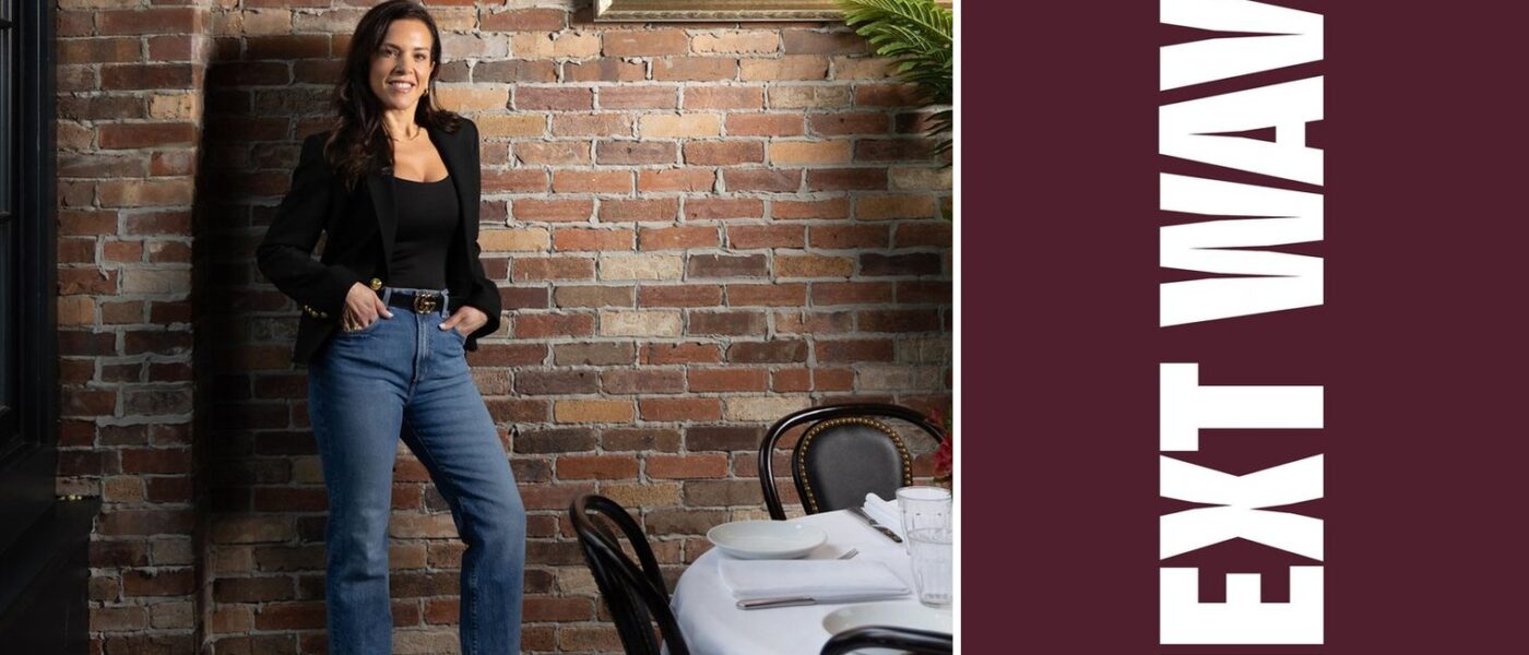 Daniella Mammola stands beside a set dining table at Daniella’s on Cabot beside vertical Next Wave lettering on maroon.