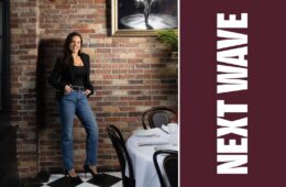 Daniella Mammola stands beside a set dining table at Daniella’s on Cabot beside vertical Next Wave lettering on maroon.