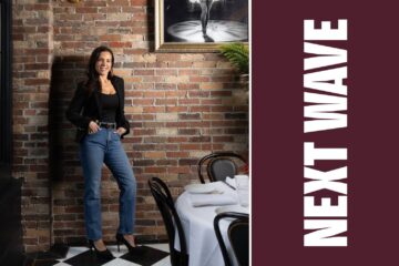Daniella Mammola stands beside a set dining table at Daniella’s on Cabot beside vertical Next Wave lettering on maroon.