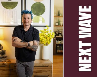 Jason Santos stands with his arms crossed in a restaurant beside vertical Next Wave lettering on maroon.