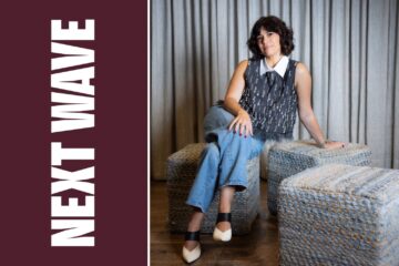 Jordan Becker sits on a woven chair at INTERLOCKS in Newburyport beside vertical Next Wave lettering on maroon.