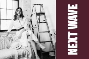 Kelsy Stromski sits near a ladder and tall windows in a black-and-white portrait beside vertical Next Wave lettering on maroon.