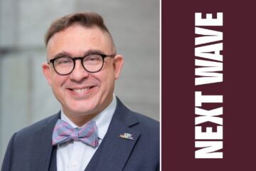 Kurt Steinberg smiles while wearing glasses, a bow tie, and a dark suit beside vertical Next Wave lettering on maroon.