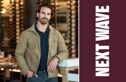 Max Faro leans against a counter with rows of wine bottles behind him beside vertical Next Wave lettering on maroon.
