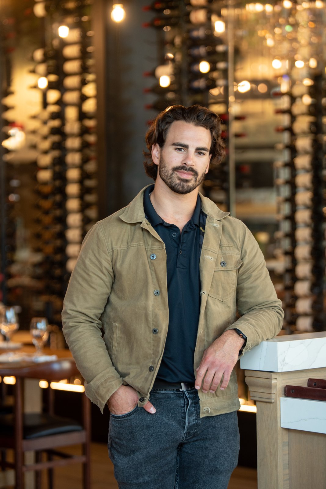 Max Faro leans against a counter with rows of wine bottles behind him.