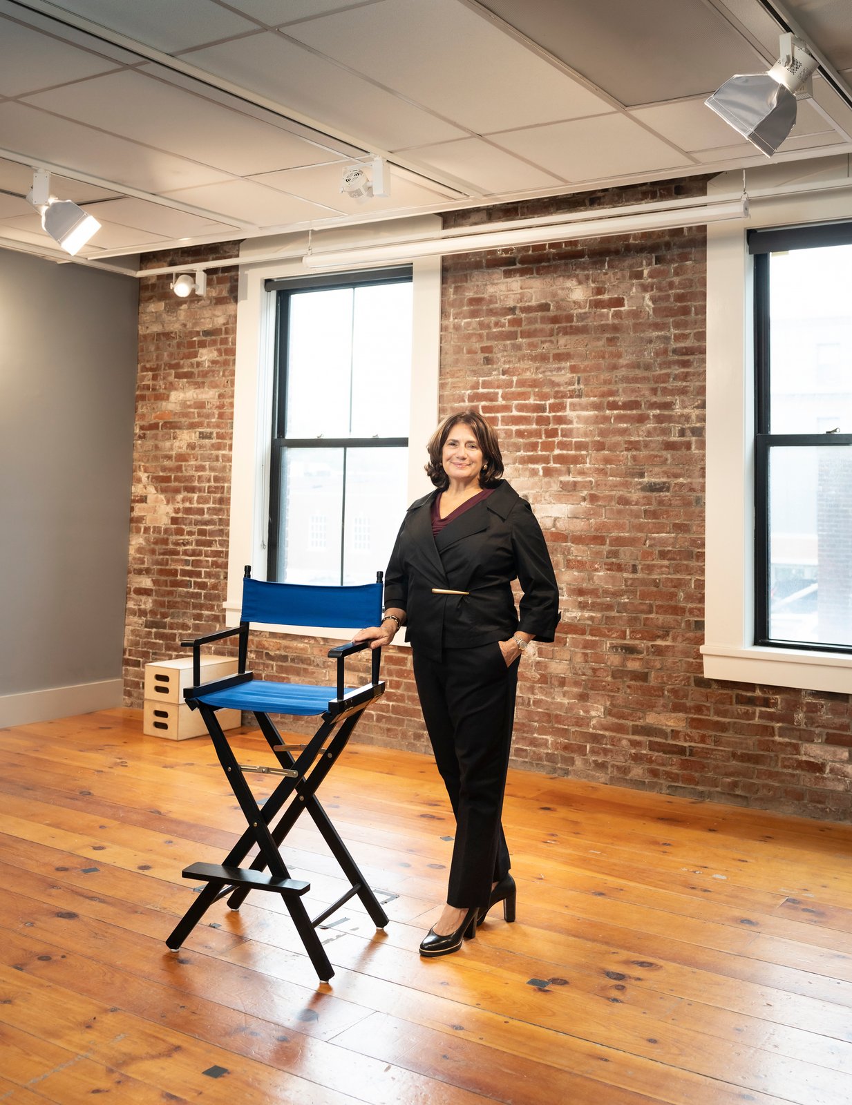 Meg Montagnino-Jarrett stands beside a blue director’s chair in a brick-walled room.