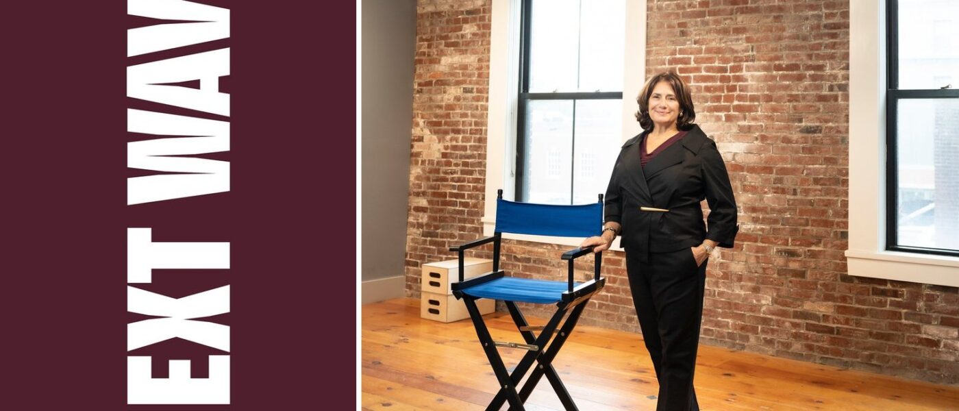 Meg Montagnino-Jarrett stands beside a blue director’s chair in a brick-walled room beside vertical Next Wave lettering on maroon.