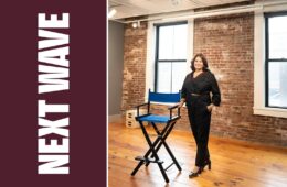 Meg Montagnino-Jarrett stands beside a blue director’s chair in a brick-walled room beside vertical Next Wave lettering on maroon.