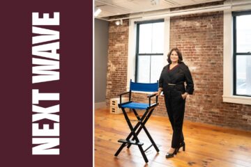 Meg Montagnino-Jarrett stands beside a blue director’s chair in a brick-walled room beside vertical Next Wave lettering on maroon.