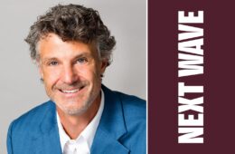 Stratton Lloyd smiles in a blue jacket and white shirt beside vertical Next Wave lettering on maroon.