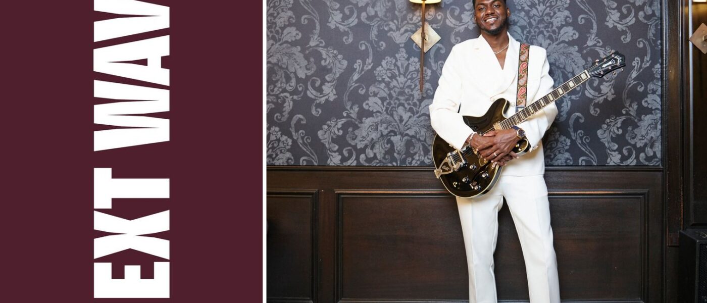 William Nelson Jr. holds an electric guitar while wearing a white suit beside vertical Next Wave lettering on maroon.
