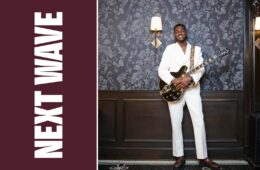 William Nelson Jr. holds an electric guitar while wearing a white suit beside vertical Next Wave lettering on maroon.