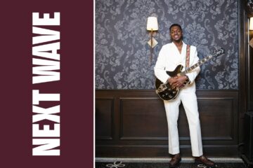 William Nelson Jr. holds an electric guitar while wearing a white suit beside vertical Next Wave lettering on maroon.